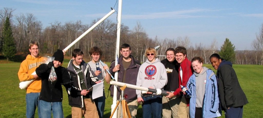 Ten students in the field posing for a photograph with potato guns after measuring the physics after potatoes are projected.