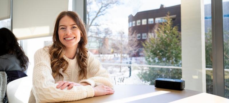 A computer science and psychology student smiles from her table in the Library with Michigan Tech's College of Computing in the background.