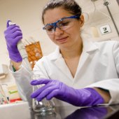 A woman in gloves and goggles pippets a liquid into a flask.