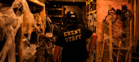 Person wearing an Event Staff shirt walking through a dark area with shelves covered in fake spiders and webs.