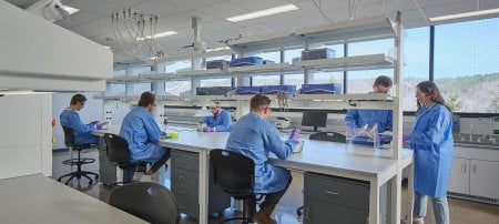 Six students in blue lab coats, purple gloves, and eye protection handle various medical equipment at a lab table.