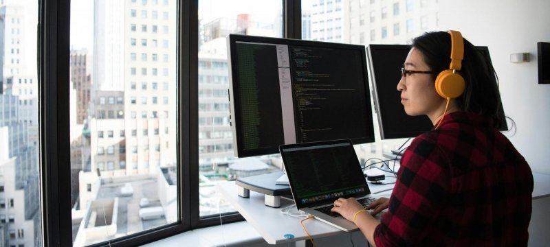 A female student works on a computer while sitting at a window in front of an urban setting.